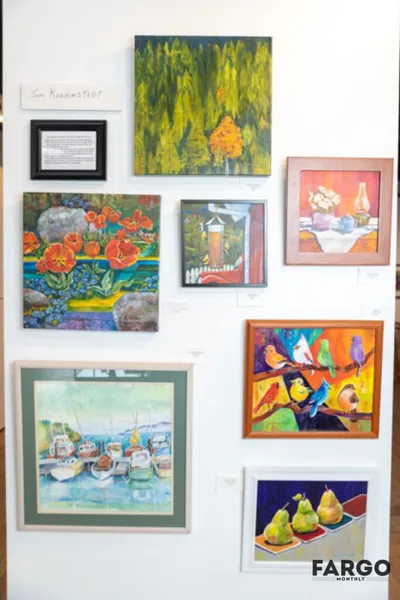Gallery wall displaying colorful paintings including florals, birds on a branch, boats, and pears