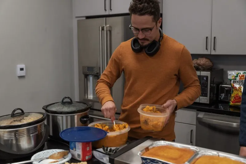 Man in orange sweater with headphones around neck serving food from containers at a kitchen counter with slow cookers nearby