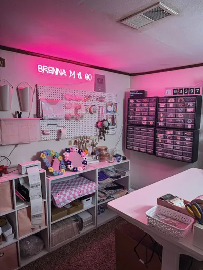 Craft room bathed in pink neon light with a pegboard, shelving, and a neon sign reading Brenna M & Co