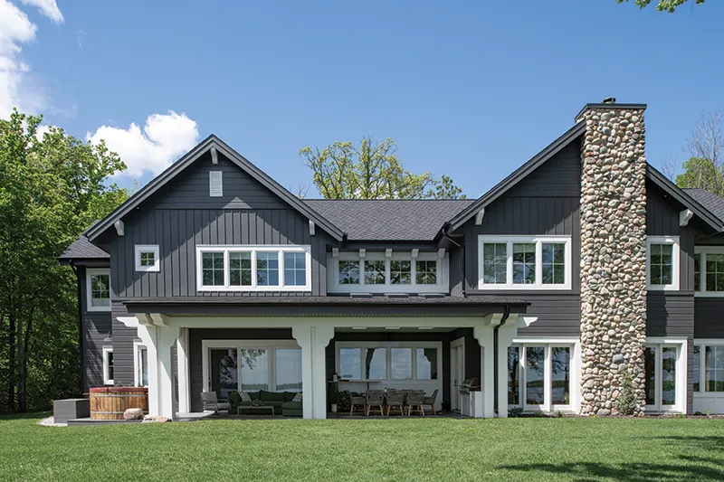 Dark gray two-story craftsman home with stone chimney, covered patio, and manicured lawn