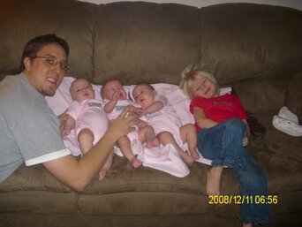 Man on a brown sofa holding three newborns in pink while a toddler rests beside them