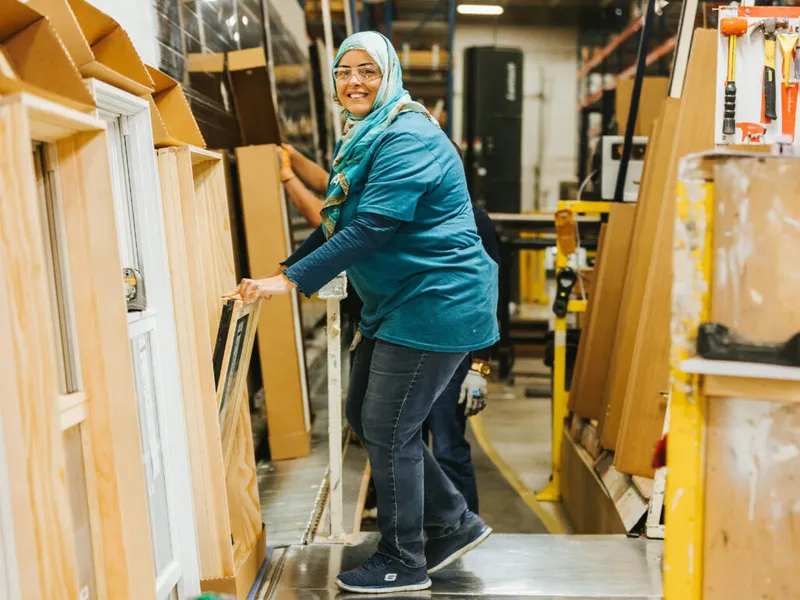 Smiling woman in teal hijab and safety glasses handling door frames in a manufacturing facility
