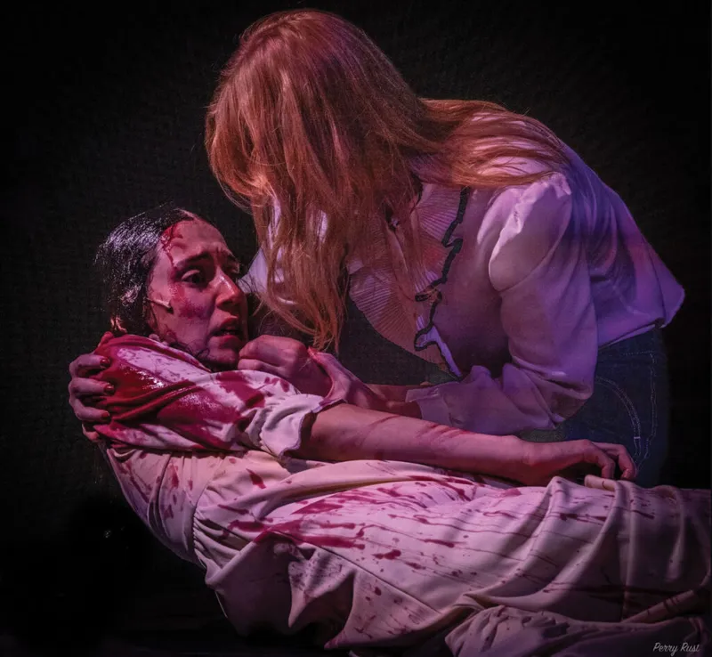 Two actors in a dramatic stage scene, one bloodied and lying down, the other leaning over them