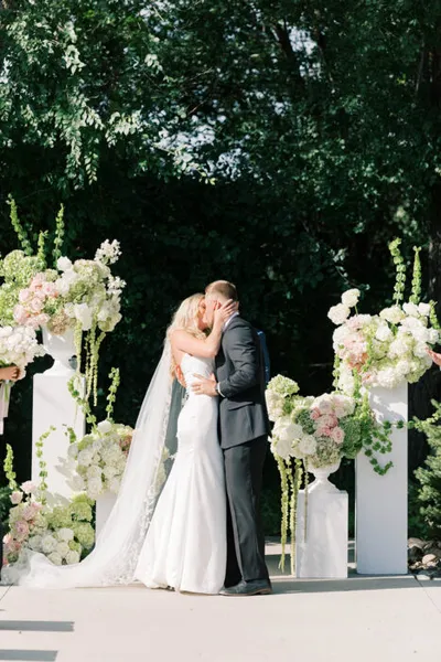 Bride and groom sharing first kiss beneath white pedestals adorned with pink and white floral arrangements outdoors
