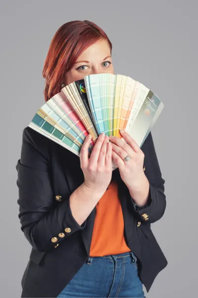 Red-haired woman in black blazer holding a fan of paint swatches over her face