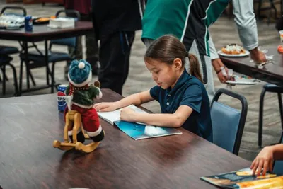 Young girl writing in a notebook at a table beside a small holiday elf decoration