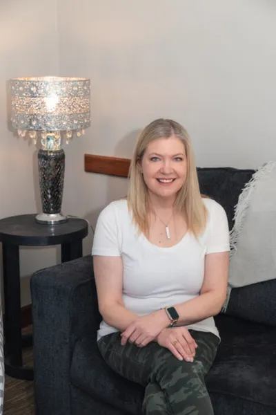 Smiling blonde woman in a white tee and camo pants seated on a dark sofa beside a decorative lamp