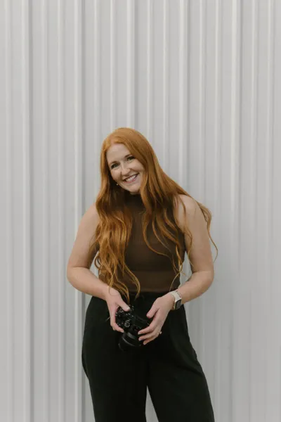 Smiling woman with long red hair holding a camera, wearing a brown sleeveless top and black pants