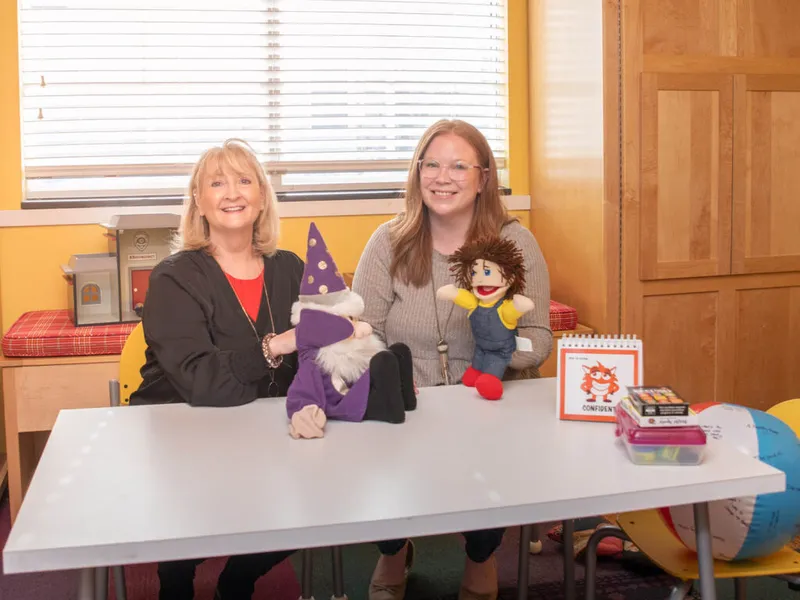 Two smiling women seated at a table holding hand puppets in a colorful classroom setting