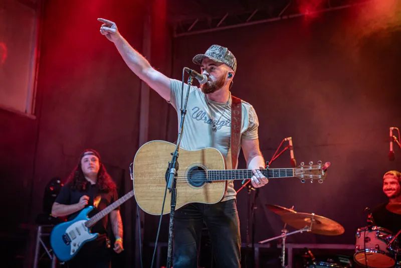 Bearded country singer in a cap and Wrangler tee points skyward while holding an acoustic guitar on a red-lit stage