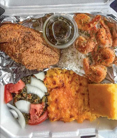Styrofoam takeout box with fried fish, breaded shrimp, rice, mac and cheese, greens, cornbread, and tomatoes