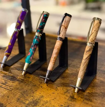 Four handcrafted pens on black stands atop a wooden table, featuring colorful resin and natural wood barrels