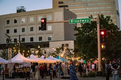Crowd gathered at an outdoor street market under string lights near a Broadway street sign at dusk