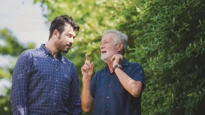 Younger man and older man with white beard talking outdoors near green hedges