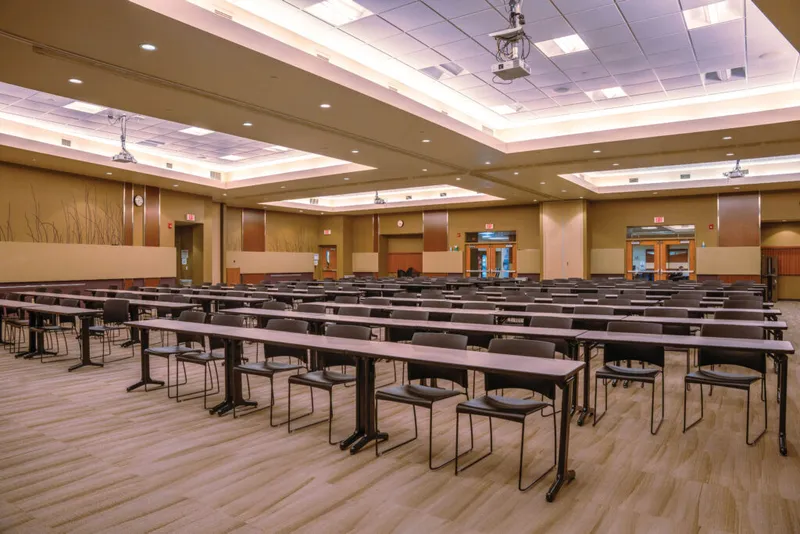 Large empty conference room with rows of folding tables and chairs beneath recessed ceiling lights and ceiling-mounted