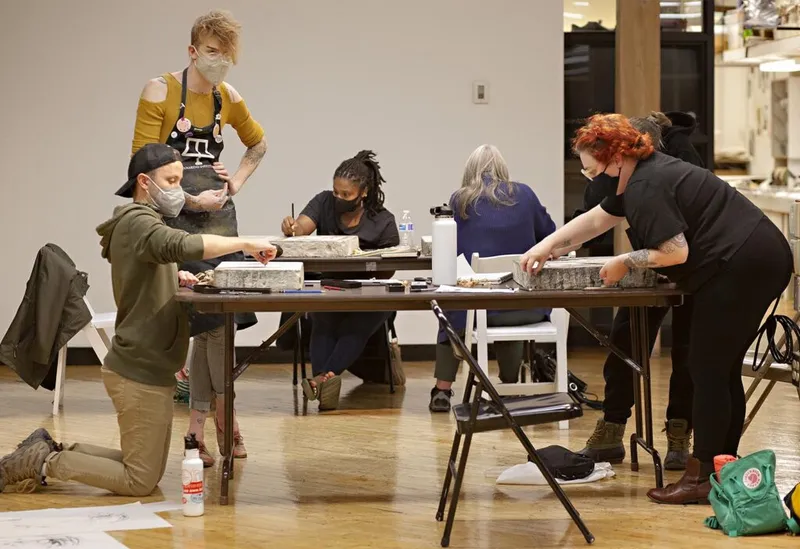 Five masked participants work around folding tables during an art workshop on a wood floor