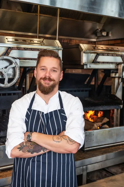 Tattooed chef in striped apron stands arms crossed before a wood-fired grill