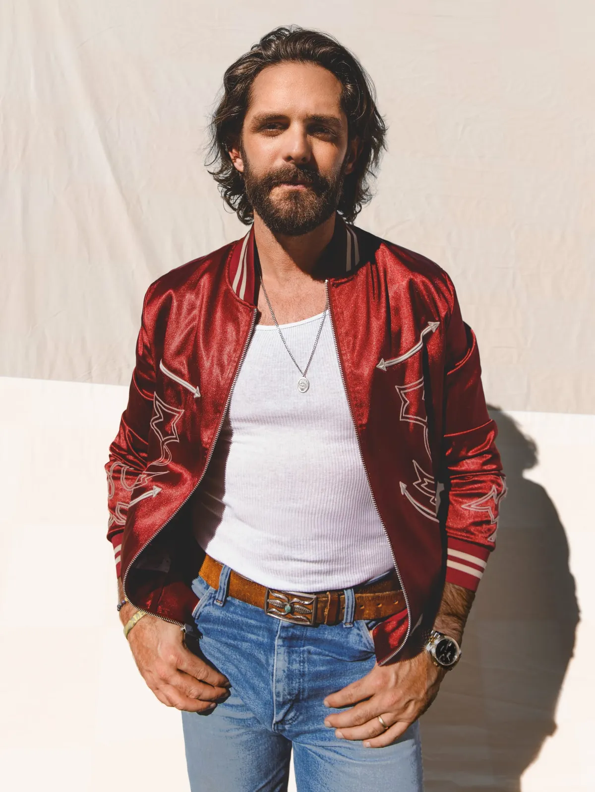 Bearded man with wavy dark hair in a red satin bomber jacket, white tank, and jeans