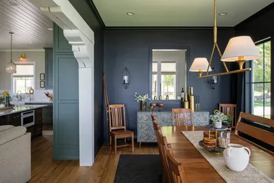 Open dining room with dark navy walls, wood table, and brass chandelier beside a green-accented kitchen