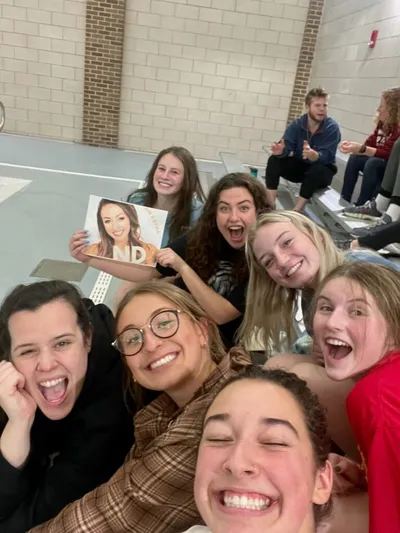 Group of laughing young women taking a selfie indoors, one holding a photo with 'ND' text