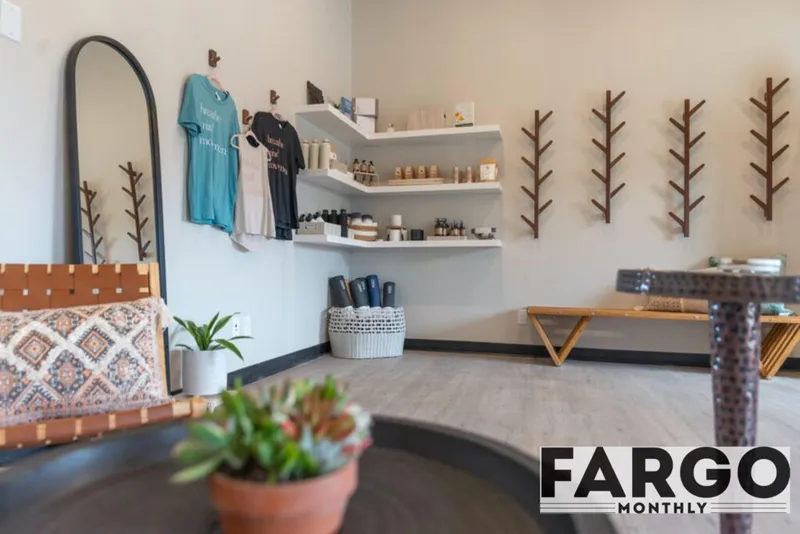 Bright yoga studio retail area with hanging tees, product shelves, tree-branch wall hooks, and a leather chair