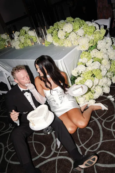 Bride and groom sitting on floor with small white cake, surrounded by white hydrangea arrangements