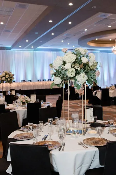 Elegant ballroom reception with tall white floral centerpiece over a round table set with gold chargers