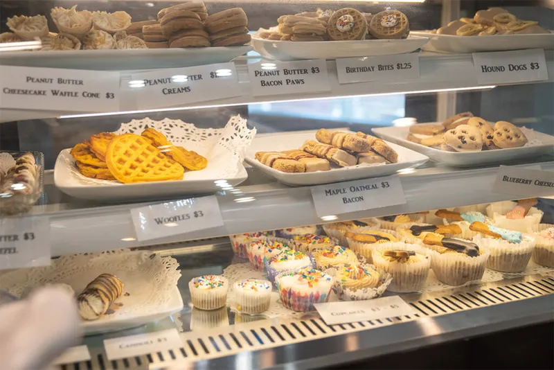 Glass bakery display case with shelves of dog treats including waffle-shaped woofles, cupcakes, and cookies with handwritten