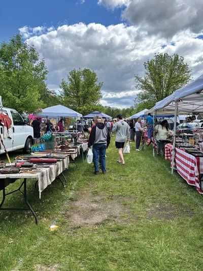 Shoppers browse vendor tables at an outdoor flea market under blue canopy tents