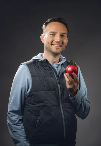 Smiling man in blue hoodie and dark vest holding a red apple