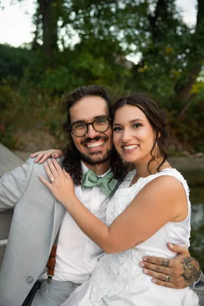 Smiling bride in white lace dress embraces groom in grey suit and green bow tie outdoors