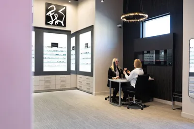 Two women seated at a small table inside a modern optical boutique with backlit eyeglass displays