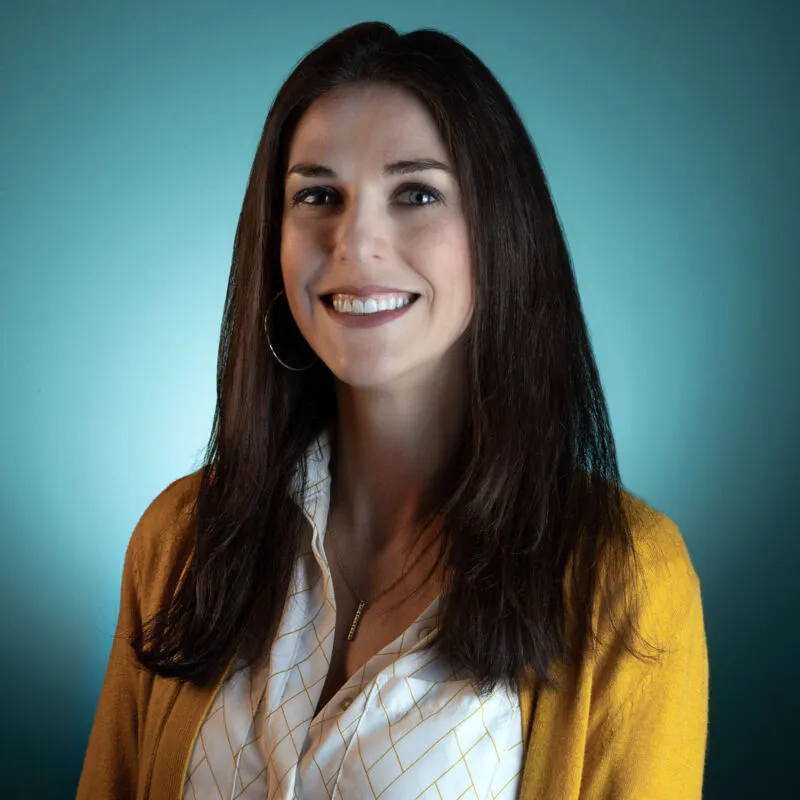 Smiling woman with long dark hair wearing a yellow cardigan over a patterned blouse against a teal background
