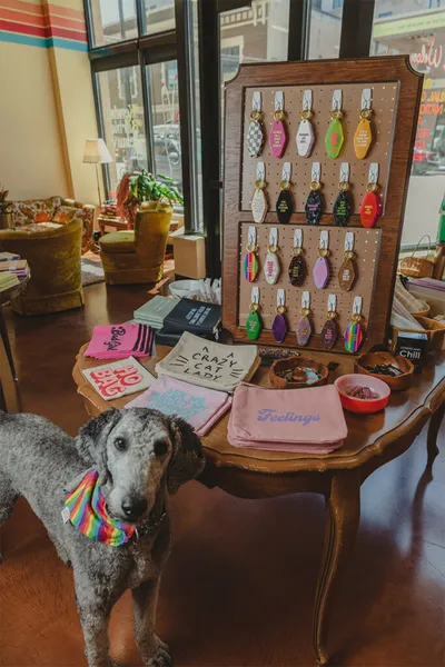 Gray dog with rainbow bandana stands beside a shop table displaying keychains and novelty pouches