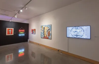 Gallery wall displaying neon 'Native Lives Matter' sign and painted canvases beside a mounted video screen