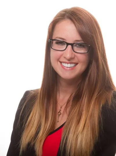 Smiling woman with long auburn hair, black-rimmed glasses, and a black blazer over a red top