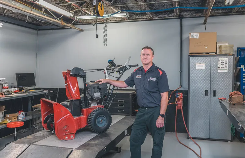 A mechanic in a dark uniform stands beside a red snowblower on a workshop lift