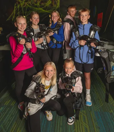 Seven kids in laser tag vests holding blasters pose together in a dimly lit arena