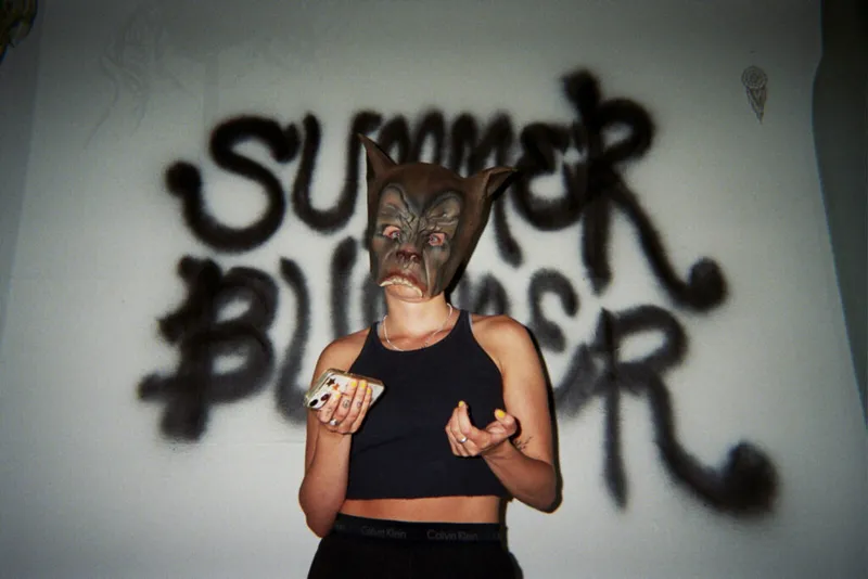 Person in a wolf mask holding a phone, standing before 'Summer Bummer' graffiti on a white wall