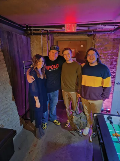 Four people posing together in a dimly lit basement venue with purple lighting and an exit sign