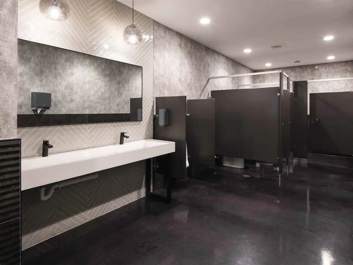 Modern public restroom with white trough sink, black faucets, and dark stall partitions