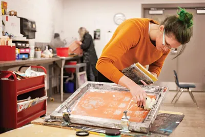 Woman in orange sweater screen printing at a large mesh frame in an art studio
