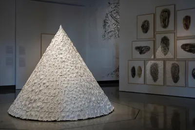 Large white textured cone sculpture in a gallery with framed artworks on the wall behind