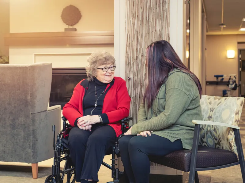 Elderly woman in a wheelchair smiling while chatting with a younger woman in a warmly lit lobby