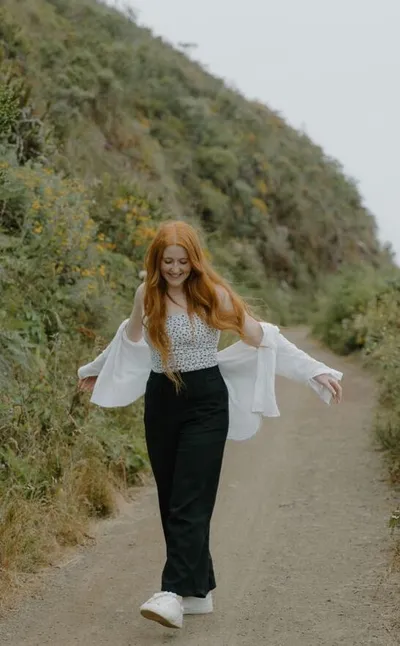 Smiling woman with long red hair walks a coastal trail in a white blouse and black wide-leg pants