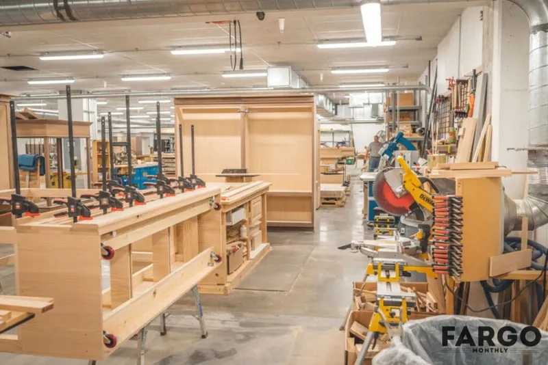 Woodworking shop with clamp-lined workbenches, a miter saw, and unfinished wood cabinetry under fluorescent lights