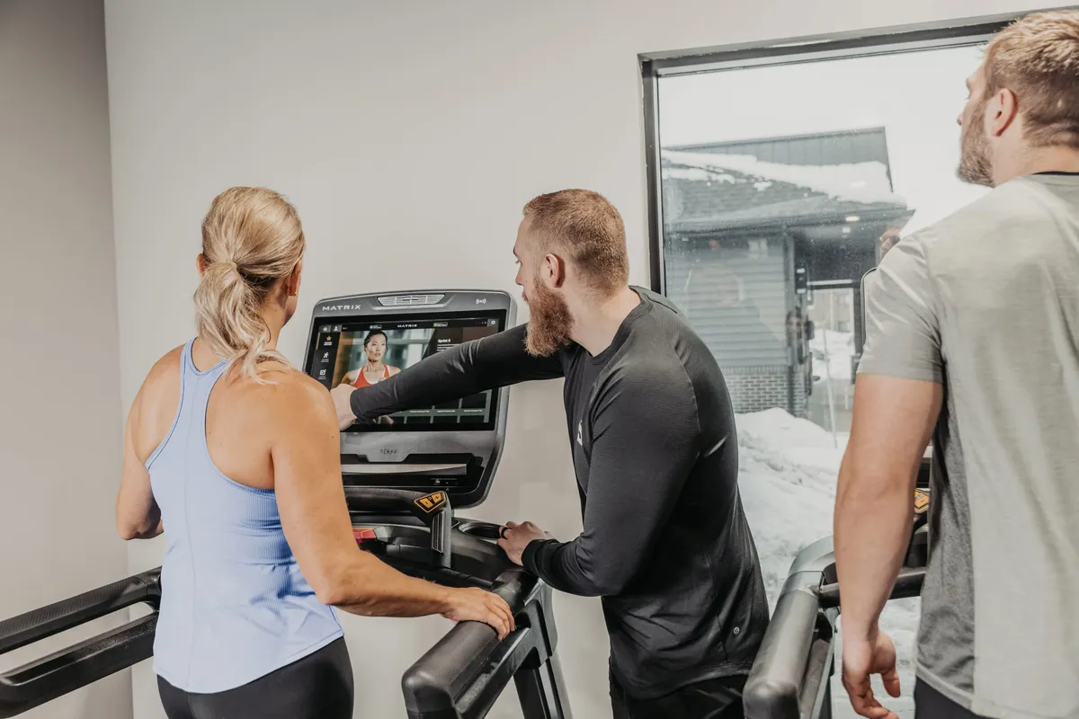 Personal trainer pointing at a Matrix treadmill touchscreen while explaining features to two gym clients