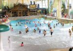 Crowded indoor wave pool with swimmers beneath a timber-framed roof