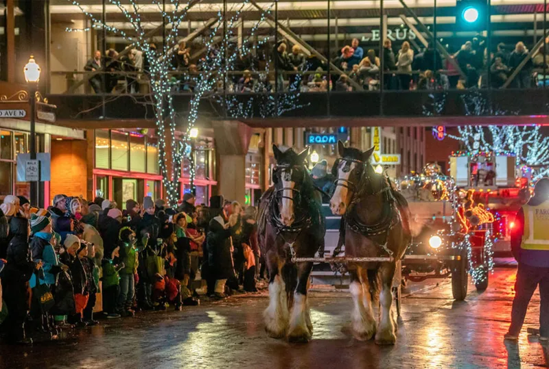 Two draft horses pulling a carriage past holiday-lit streets lined with bundled spectators at night