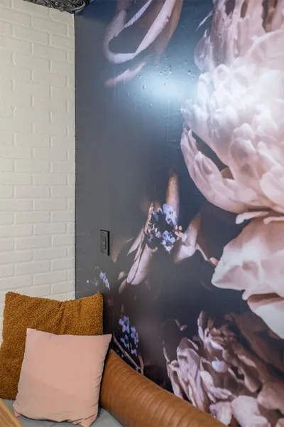 Leather sofa with pink and mustard cushions beside a dark floral mural and white brick wall